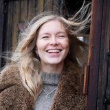 Smiling blonde woman in a brown shearling coat outdoors beside a wooden doorway.